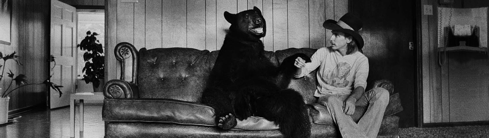 Black bear sitting on a couch with a man in a hat in the 70s